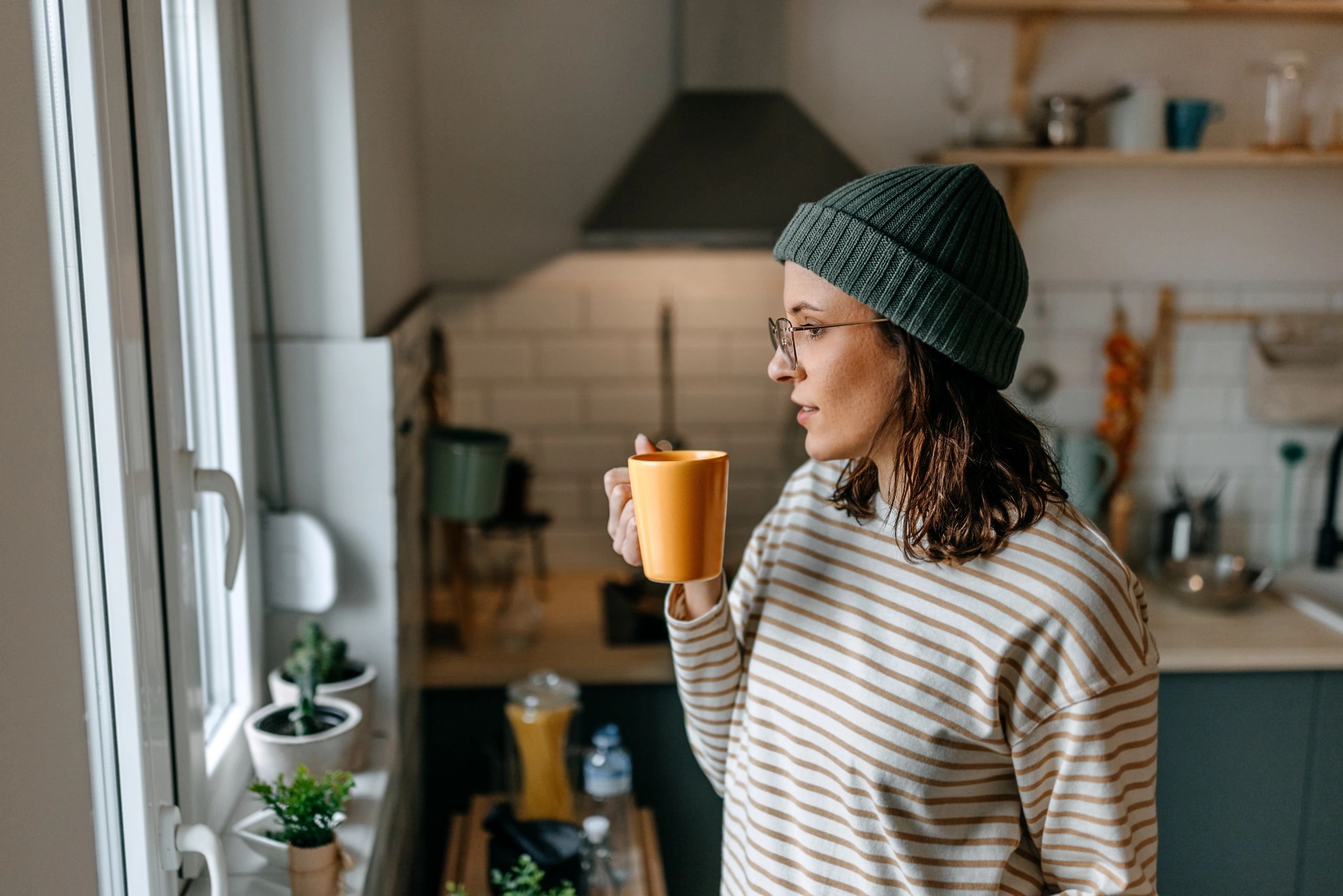 Woman with coffee cup looking through window at home