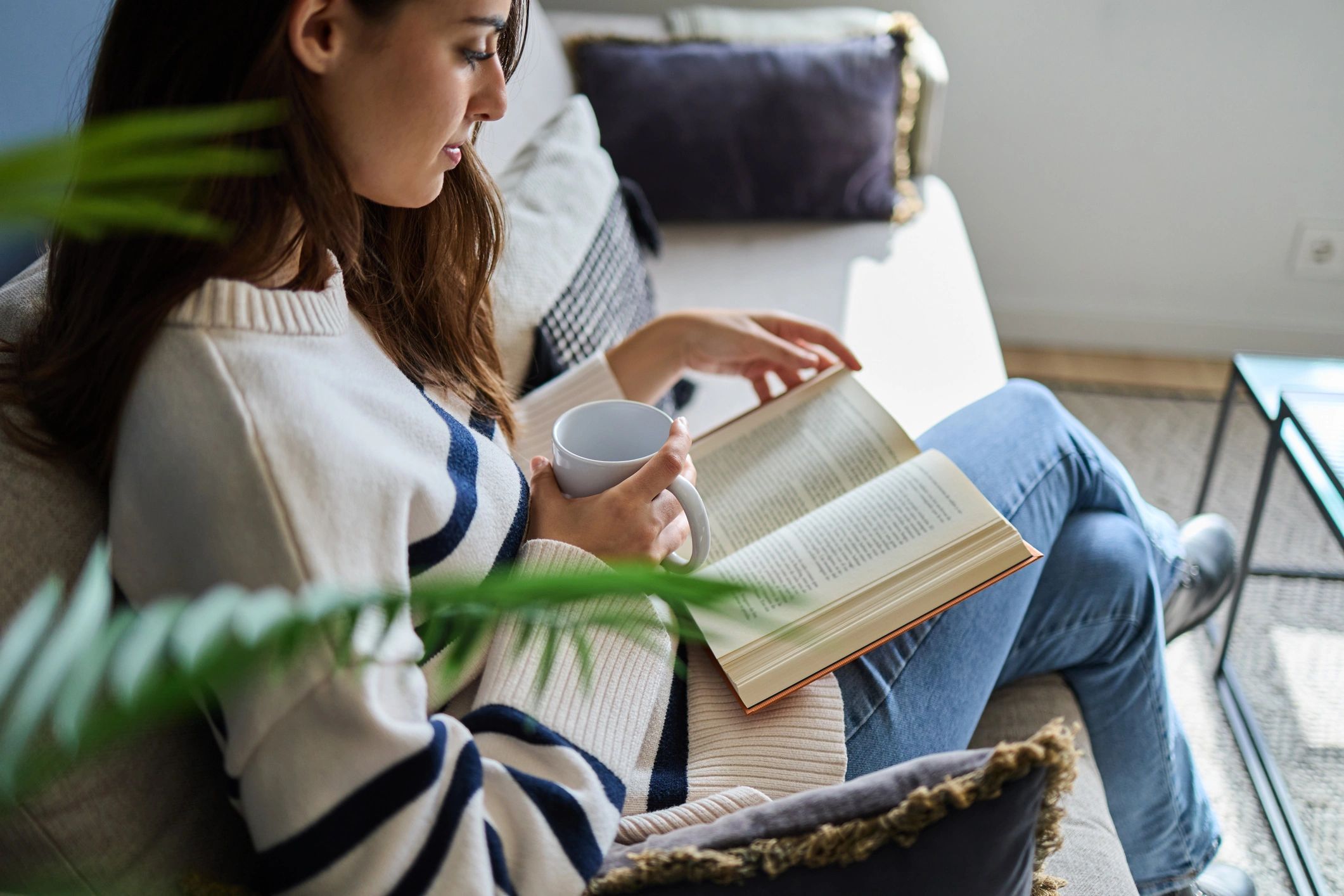 Overhead view of a woman relaxing on a sofa with a book and warm drink